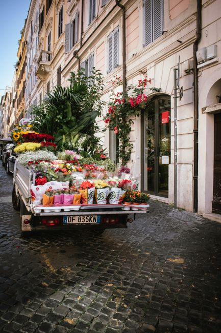 A flower delivery truck parked on a cobblestone street in front of a historical building with tall windows and decorative exterior details. The truck's flatbed is loaded with an array of freshly cut f